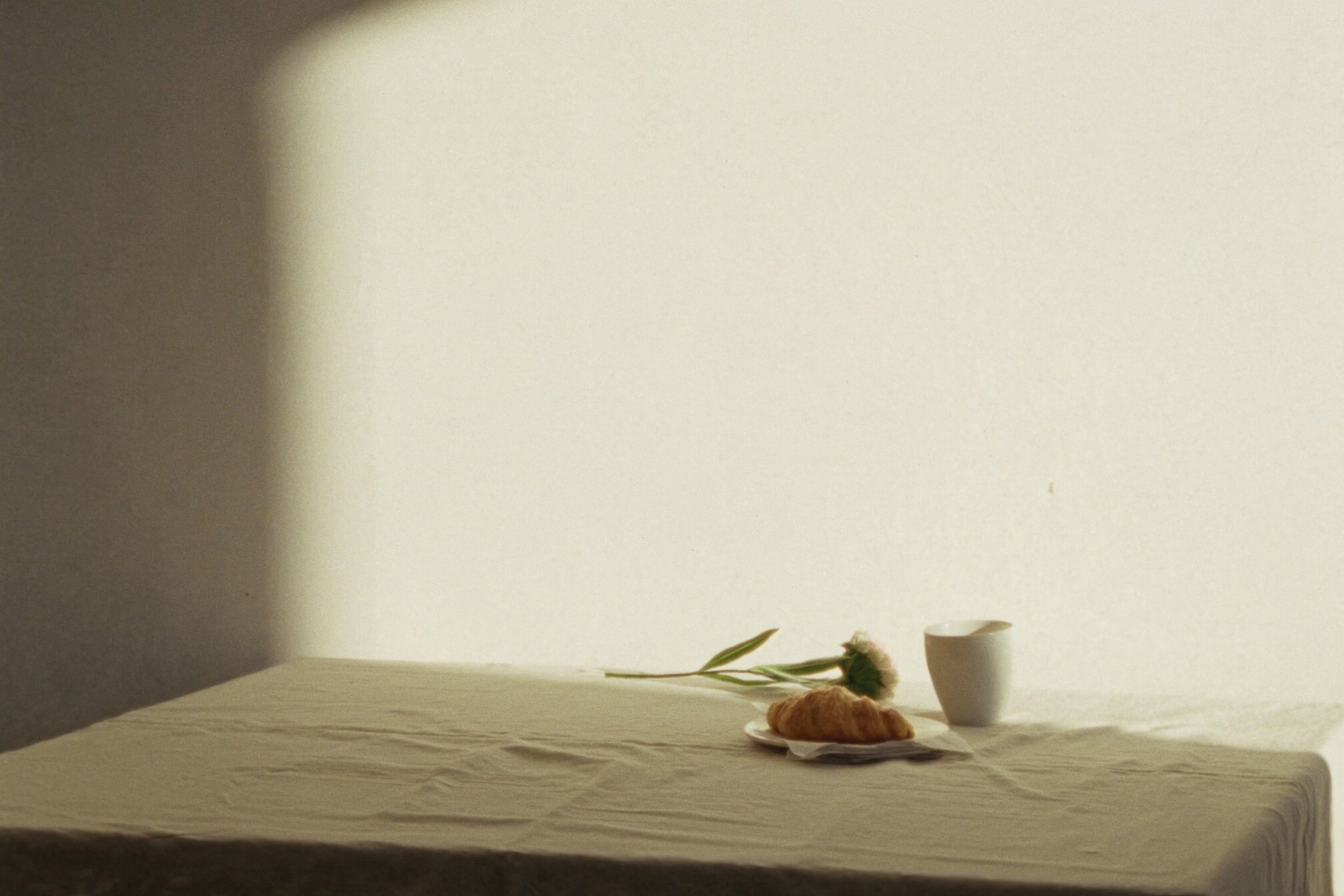 A sparse breakfast table with linen, a cup, pastry, and a single flower in soft light.
