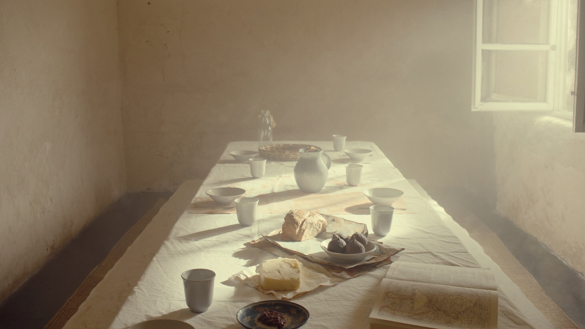 A long quiet breakfast table in a sunlit room with linen, cups, bread, and an open window.